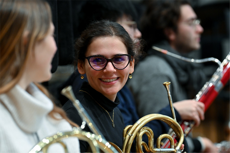 A photo of a student, holding a French horn, smiling at a student, sitting in an orchestra.